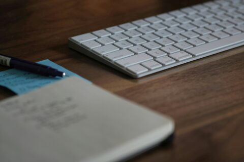 Foto von Tastatur im Fokus und davor unscharf Stift und Notizbuch auf einem Tisch